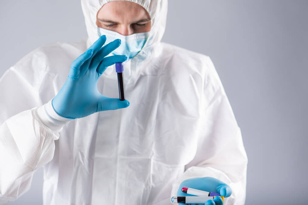 Um virologista em uma máscara médica e roupas de proteção possui um tubo de teste com uma amostra de sangue para testes de coronavírus. Pandemia. Síndrome respiratória, pânico, experiências, pesquisa
 - Foto, Imagem
