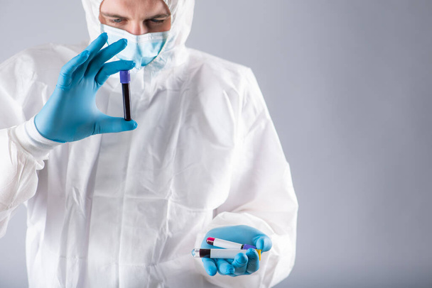 Virolog v lékařské masce a ochranném oděvu má zkumavku se vzorkem krve pro testování koronaviru. Pandemie. Respirační syndrom, panika, zkušenosti, výzkum - Fotografie, Obrázek