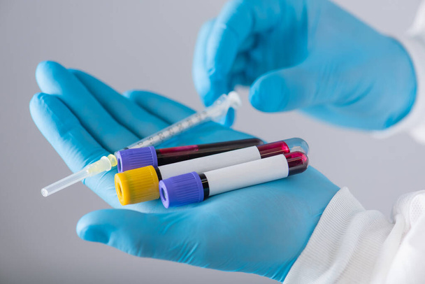 virolog v ochranném obleku s dýchací maskou a modrými rukavicemi drží injekční stříkačku a krevní testy ve zkumavkách - Fotografie, Obrázek