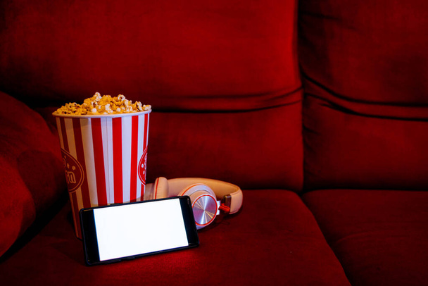 mobile phone with empty white bright screen with popcorn bucket on the red sofa. Concept of streaming TV on internet phone. - Photo, Image