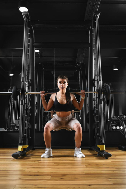 A girl is training her legs in the gym with a barbell. Sport lifestyle, keep fit, fitness motivation. dark background, copy space - Photo, Image