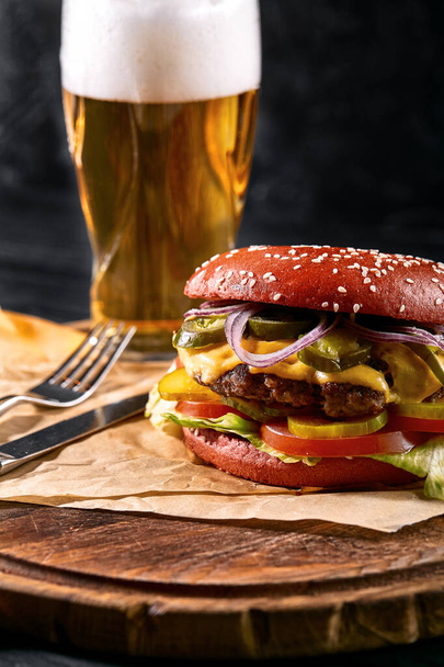 Delicious hot spicy black burger with chili pepper and glass of beer on cutting board on white wood table - Photo, image