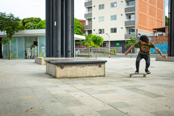Medellin, Antiokvia / Kolumbie - 10. října 2019: Hispánec s dlouhými hnědými vlasy dělá skateboarding Back Side Smith Grind Trick na kovové lavičce ve veřejném parku - Fotografie, Obrázek