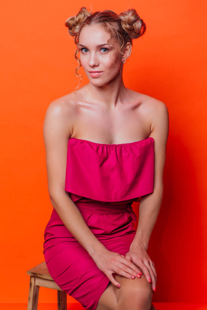 The girl in a red dress on a orange background in the studio. Blonde girl with two hair knots holding hands on knees, looking to the camera and smiling. - Fotografie, Obrázek