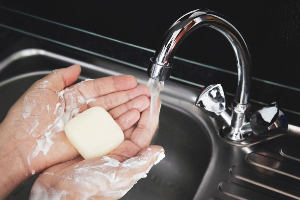 Lavarse las manos con jabón bajo agua corriente. pedazo de jabón en las palmas de los hombres bajo la corriente de agua contra el fondo de un fregadero de metal
. - Foto, Imagen