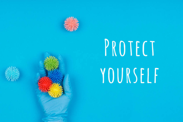 Χέρι στο γάντι μιας χρήσης με πλαστικές μπάλες ιούς στο μπλε φόντο με Προστατέψτε τον εαυτό σας διατύπωση. Επιδημικοί, εποχιακοί ιοί, κοινωνική απομόνωση, κλείσιμο, έννοια coronavirus COVID-19 - Φωτογραφία, εικόνα