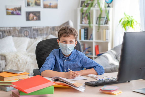 Boy in face mask with computer does his homework during a coronavirus quarantine. concept of online education. Social distance, self- isolation. Distance learning due to virus, flu, epidemic COVID-19 - 写真・画像