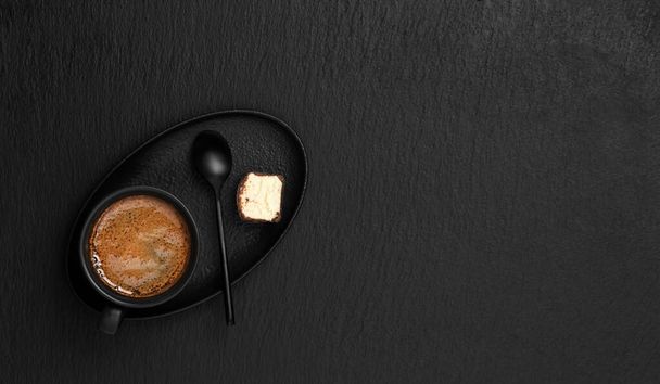Black cup with espresso, on a saucer a dessert and a spoon, black stone background. Copy space for text. View from above. Morning hours waiting for coffee. - Фото, изображение