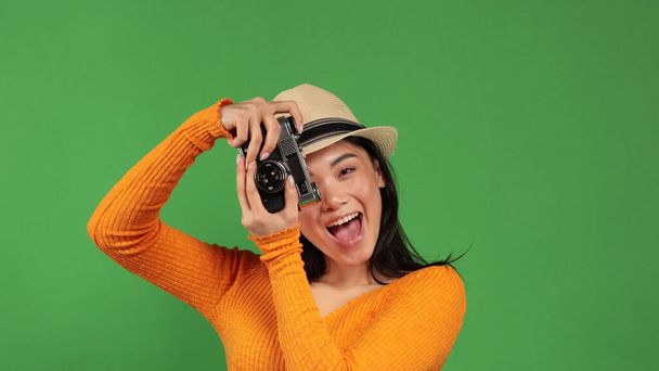 Asian woman in stylish outfit actively taking photos through a retro camera on green background. Hobbies, leisure, favorite pastime. Fun, relaxation, photoset. Female portrait - Фото, зображення
