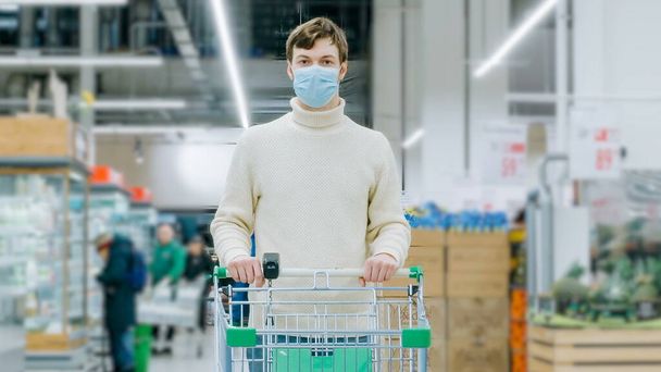 Tıbbi maskeli bir adam elinde yemek arabası olan bir süpermarkette duruyor. - Fotoğraf, Görsel