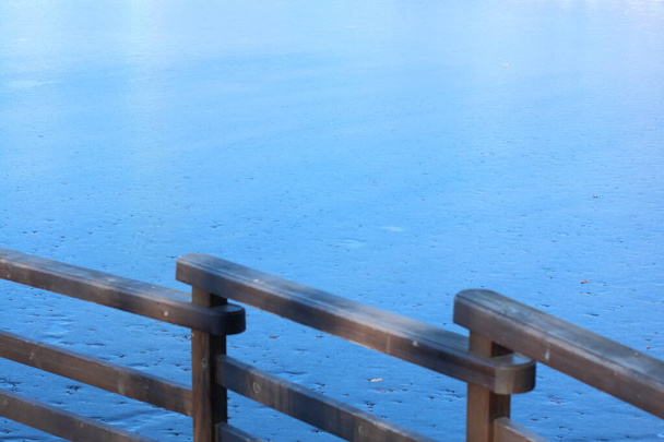 παγωμένη λίμνη σε μια καθαρή χειμωνιάτικη μέρα, καλυμμένη με μπλε πάγο, σε πρώτο πλάνο μπορείτε να δείτε το ξύλινο κιγκλίδωμα της προβλήτας - Φωτογραφία, εικόνα