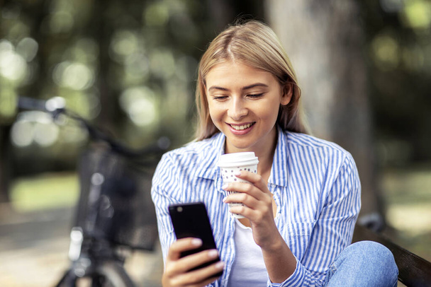 Блондинка молодая женщина сидит на скамейке, смотрит на мобильный телефон и пьет кофе, чтобы пойти в парк с велосипедом на заднем плане - Фото, изображение