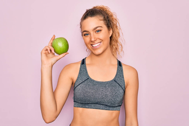 Nuori kaunis blondi urheilullinen nainen sinisilmäinen tilalla terve vihreä omena hedelmiä onnellinen kasvot seisoo ja hymyilee luottavainen hymy osoittaa hampaat
 - Valokuva, kuva