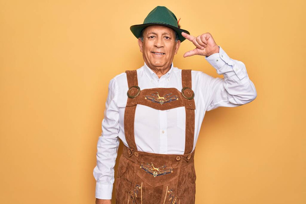Senior grey-haired man wearing german traditional octoberfest suit over yellow background smiling and confident gesturing with hand doing small size sign with fingers looking and the camera. Measure concept. - Foto, Bild