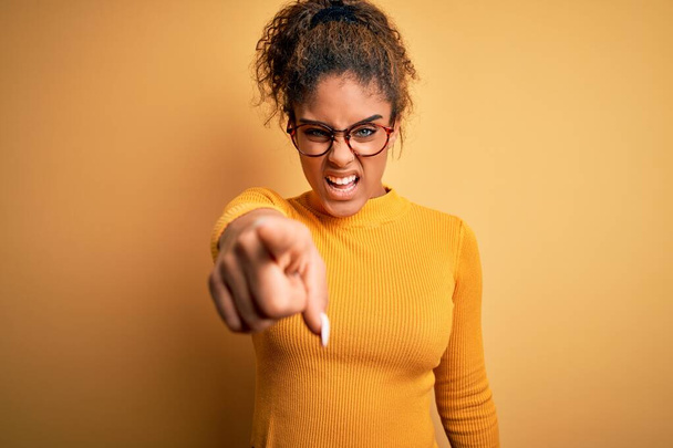 Jeune belle fille afro-américaine portant un pull et des lunettes sur fond jaune pointant mécontent et frustré à la caméra, en colère et furieux contre vous
 - Photo, image