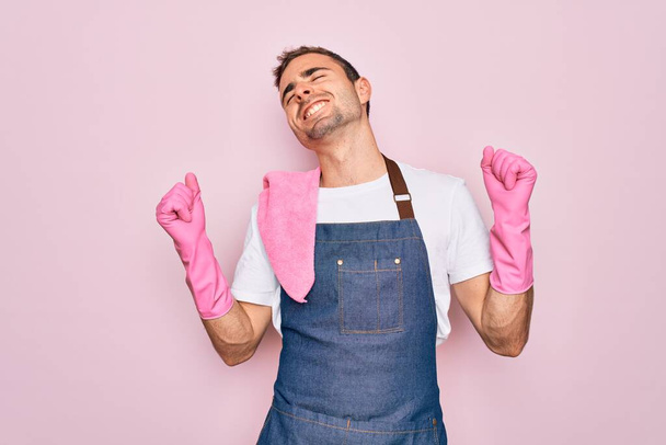 Junger Putzmann mit blauen Augen beim Putzen mit Schürze und Handschuhen über rosa Hintergrund sehr glücklich und aufgeregt dabei Siegergeste mit erhobenen Armen, lächelnd und schreiend nach Erfolg. Festkonzept. - Foto, Bild