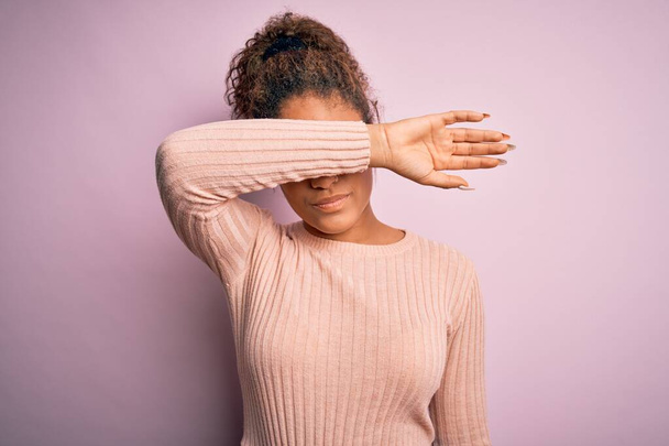 Mladý krásný africký americký dívka na sobě ležérní svetr stojící nad růžovým pozadím zakrývající oči s paží, vypadající vážné a smutné. Slepý, skrývající a odmítavý koncept - Fotografie, Obrázek