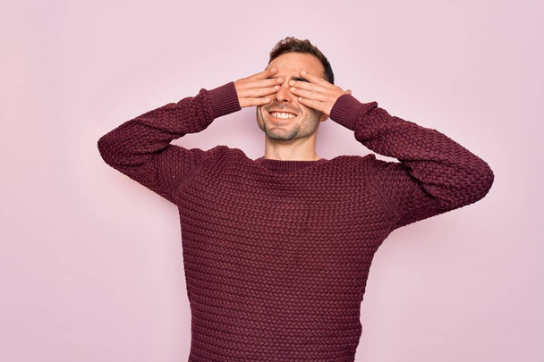 Young handsome man with blue eyes wearing casual sweater standing over pink background covering eyes with hands smiling cheerful and funny. Blind concept. - Photo, Image