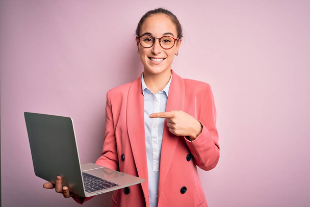 Młoda, piękna bizneswoman nosząca okulary pracująca na laptopie na różowym tle bardzo szczęśliwa wskazując ręką i palcem - Zdjęcie, obraz
