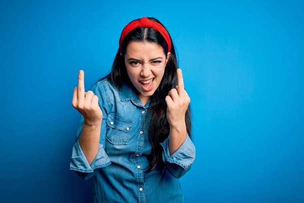 Young brunette woman wearing casual denim shirt over blue isolated background Showing middle finger doing fuck you bad expression, provocation and rude attitude. Screaming excited - Fotoğraf, Görsel