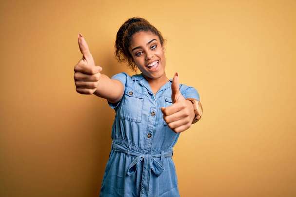 Молодая красивая африканская американка в джинсовом платье, стоящая на жёлтом фоне, одобряя позитивный жест рукой, улыбается и радуется успеху. Жест победителя
. - Фото, изображение