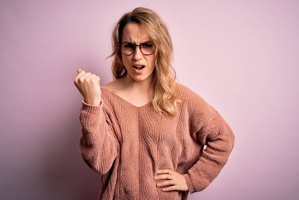 Fiatal gyönyörű szőke nő visel alkalmi pulóver és szemüveg felett rózsaszín háttér dühös és őrült emelő ököl frusztrált és dühös, miközben kiabál a harag. Düh és agresszív koncepció. - Fotó, kép