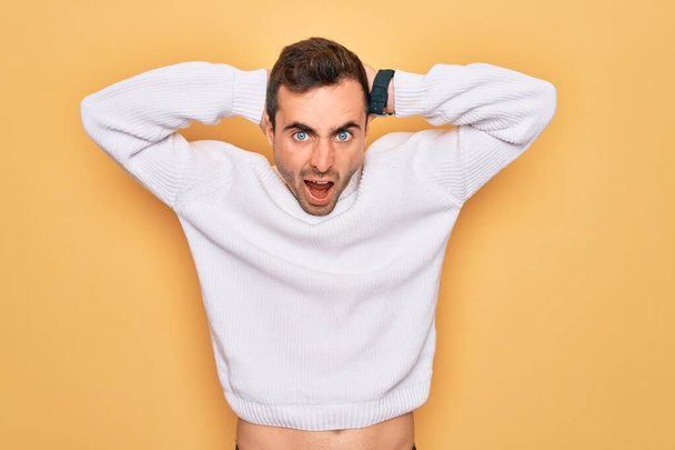 Young handsome man with blue eyes wearing casual sweater standing over yellow background Crazy and scared with hands on head, afraid and surprised of shock with open mouth - Photo, Image