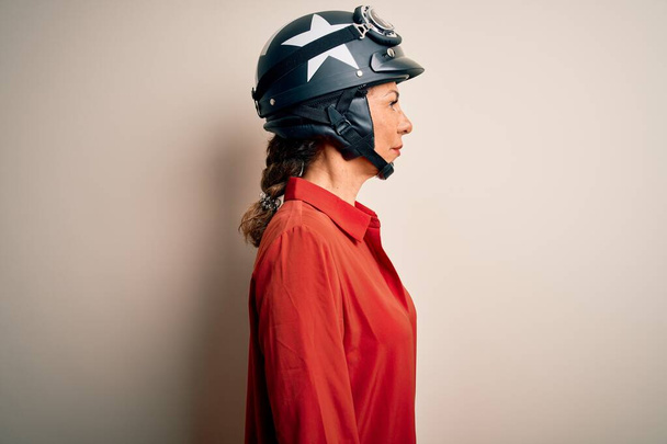Mujer motociclista de mediana edad que usa casco de motocicleta sobre un fondo blanco aislado mirando al costado, pose de perfil relajado con cara natural con sonrisa confiada
. - Foto, Imagen