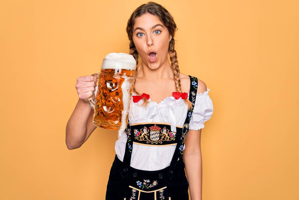 Mooie blonde Duitse vrouw met blauwe ogen in oktober jurk drinken pot bier bang in shock met een verrassing gezicht, bang en opgewonden met angst uitdrukking - Foto, afbeelding