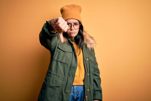 Young brunette woman wearing glasses and winter coat with hat over yellow isolated background looking unhappy and angry showing rejection and negative with thumbs down gesture. Bad expression. - Photo, Image
