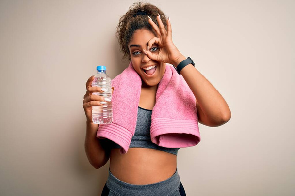 Fiatal afro-amerikai sportos nő csinál sport visel törölköző ivás üveg víz boldog arc mosolyog csinál rendben jel kéz a szemen néz keresztül ujjait - Fotó, kép