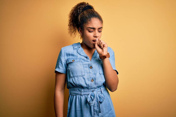 Young beautiful african american girl wearing denim dress standing over yellow background feeling unwell and coughing as symptom for cold or bronchitis. Health care concept. - Photo, Image