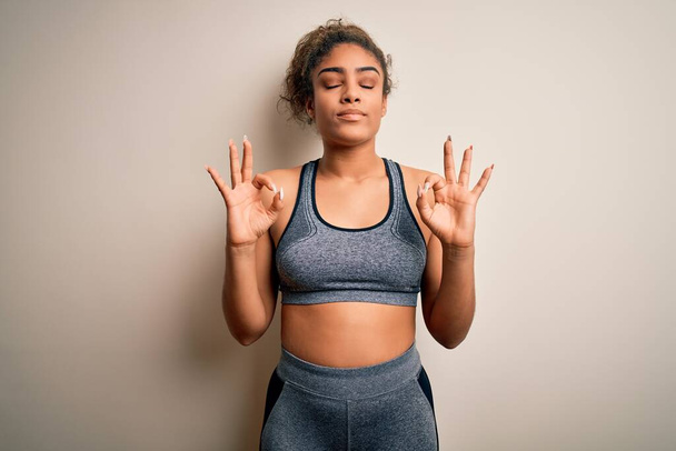 Genç Afrikalı Amerikalı sporcu kadın spor yapıyor beyaz arka planda spor kıyafetleri giyiyor rahat ve gözleri kapalı gülümsüyor parmaklarıyla meditasyon hareketi yapıyor. Yoga konsepti. - Fotoğraf, Görsel