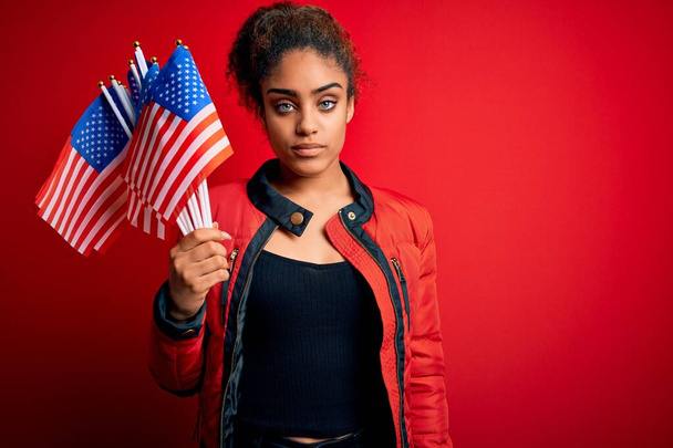 Chica patriótica afroamericana joven sosteniendo banderas de estados unidos sobre fondo rojo aislado con una expresión segura en cara inteligente pensando seriamente
 - Foto, Imagen
