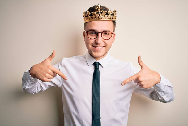 Молодой красивый кавказский бизнесмен, одетый в золотую корону на изолированном фоне, уверенно выглядящий с улыбкой на лице, указывающий на себя пальцами гордым и счастливым
. - Фото, изображение