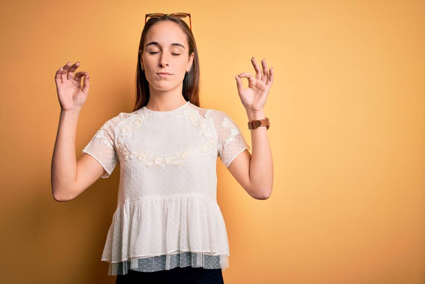 Young beautiful woman wearing casual t-shirt and sunglasses over isolated yellow background relax and smiling with eyes closed doing meditation gesture with fingers. Yoga concept. - Photo, Image