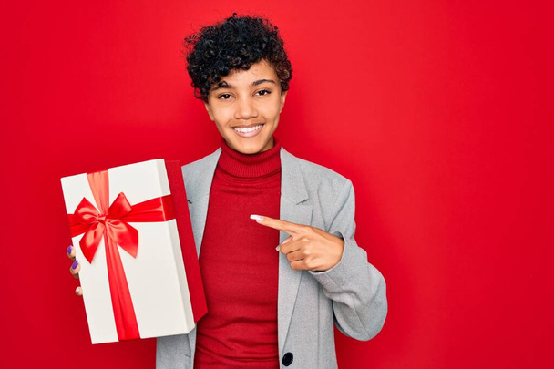 Красивая африканская американская афро-бизнес-женщина держит подарок на изолированном красном фоне очень счастливы указывая рукой и пальцем
 - Фото, изображение