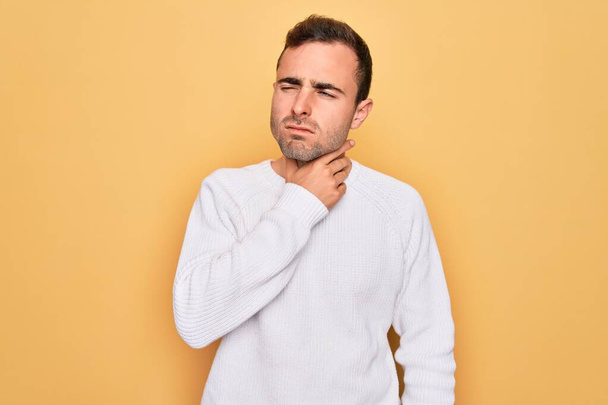 Young handsome man with blue eyes wearing casual sweater standing over yellow background Touching painful neck, sore throat for flu, clod and infection - Фото, зображення