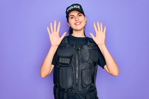 Mulher jovem polícia vestindo uniforme colete à prova de balas de segurança sobre fundo roxo mostrando e apontando para cima com os dedos número dez enquanto sorri confiante e feliz
. - Foto, Imagem