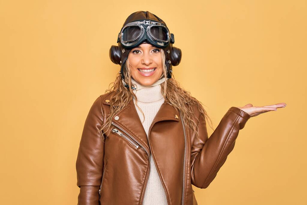 Joven hermosa aviador rubia mujer con casco piloto vintage whit gafas y chaqueta sonriente alegre presentando y señalando con la palma de la mano mirando a la cámara
. - Foto, Imagen