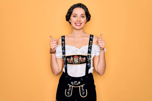 Young beautiful german woman with blue eyes wearing traditional octoberfest dress success sign doing positive gesture with hand, thumbs up smiling and happy. Cheerful expression and winner gesture. - Fotografie, Obrázek