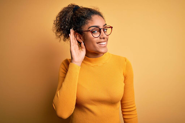 Jovem menina afro-americana bonita vestindo suéter e óculos sobre fundo amarelo sorrindo com a mão sobre o ouvido ouvindo uma audição para boatos ou fofocas. Conceito de surdez
. - Foto, Imagem