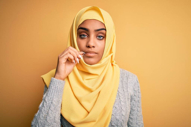 Молодая красивая африканская американская девушка в мусульманском хиджабе на изолированном желтом фоне рот и губы закрыты, как молния с пальцами. Тайное и молчаливое, табу.
 - Фото, изображение