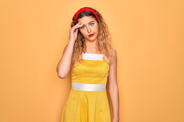 Beautiful blonde pin-up woman with blue eyes wearing diadem standing over yellow background worried and stressed about a problem with hand on forehead, nervous and anxious for crisis - Foto, Imagem