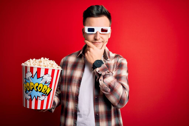 Молодий красивий кавказький чоловік носить 3d кіноокуляри і їсть попкорн, дивлячись впевнено на камеру, посміхаючись схрещеними руками і піднятою рукою на підборідді. Мислення позитивне
. - Фото, зображення