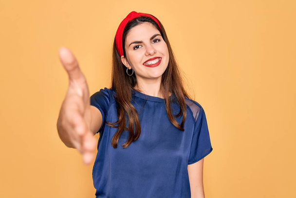 Mladá krásná brunetka žena na sobě červené rty přes žluté pozadí s úsměvem přátelské nabízí potřesení rukou jako pozdrav a přivítání. Úspěšná práce. - Fotografie, Obrázek