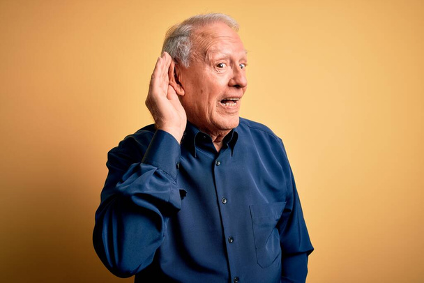 Ένας γκριζομάλλης ηλικιωμένος που φοράει μπλε πουκάμισο στέκεται πάνω από κίτρινο φόντο χαμογελώντας με το χέρι πάνω από το αυτί ακούγοντας μια ακρόαση για φήμες ή κουτσομπολιά. Έννοια κώφωση. - Φωτογραφία, εικόνα
