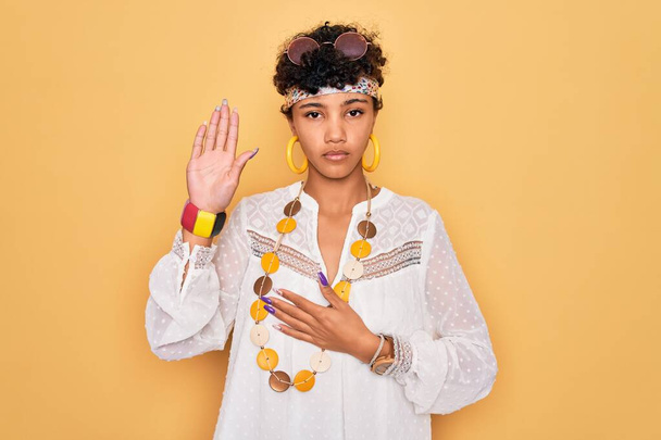 若い美しいアフリカ系アメリカ人のアフロヒッピーの女性がサングラスとアクセサリーを身に着けています胸と開いた手のひらに手で身に着けています,忠誠の誓いを作ります - 写真・画像
