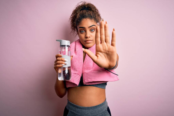 Jeune fille sportive afro-américaine faisant du sport portant une serviette bouteille d'eau à la main ouverte faisant signe d'arrêt avec expression sérieuse et confiante, geste de défense
 - Photo, image