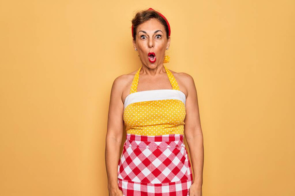 Middle age senior pin up housewife woman wearing 50s style retro dress and apron afraid and shocked with surprise expression, fear and excited face. - Photo, Image